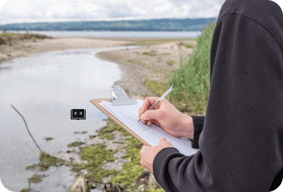 A person engaged in environmental consulting takes notes on a clipboard while observing the coastal landscape, where the serene water, lush greenery, and cloudy skies provide a picturesque backdrop.