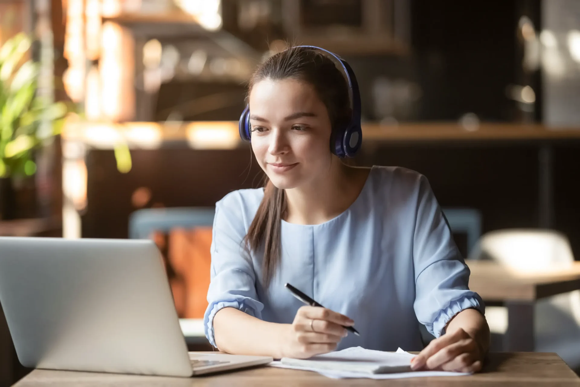 A woman wearing headphones sits at a table with a laptop, taking notes on paper, in a warmly lit room.