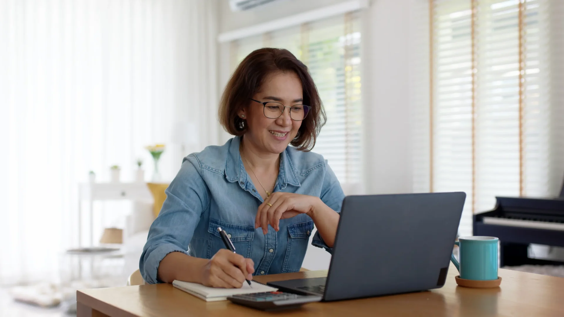 A woman in a denim shirt is sitting at a table, writing in a notebook while using a laptop. A calculator and a mug are on the table.