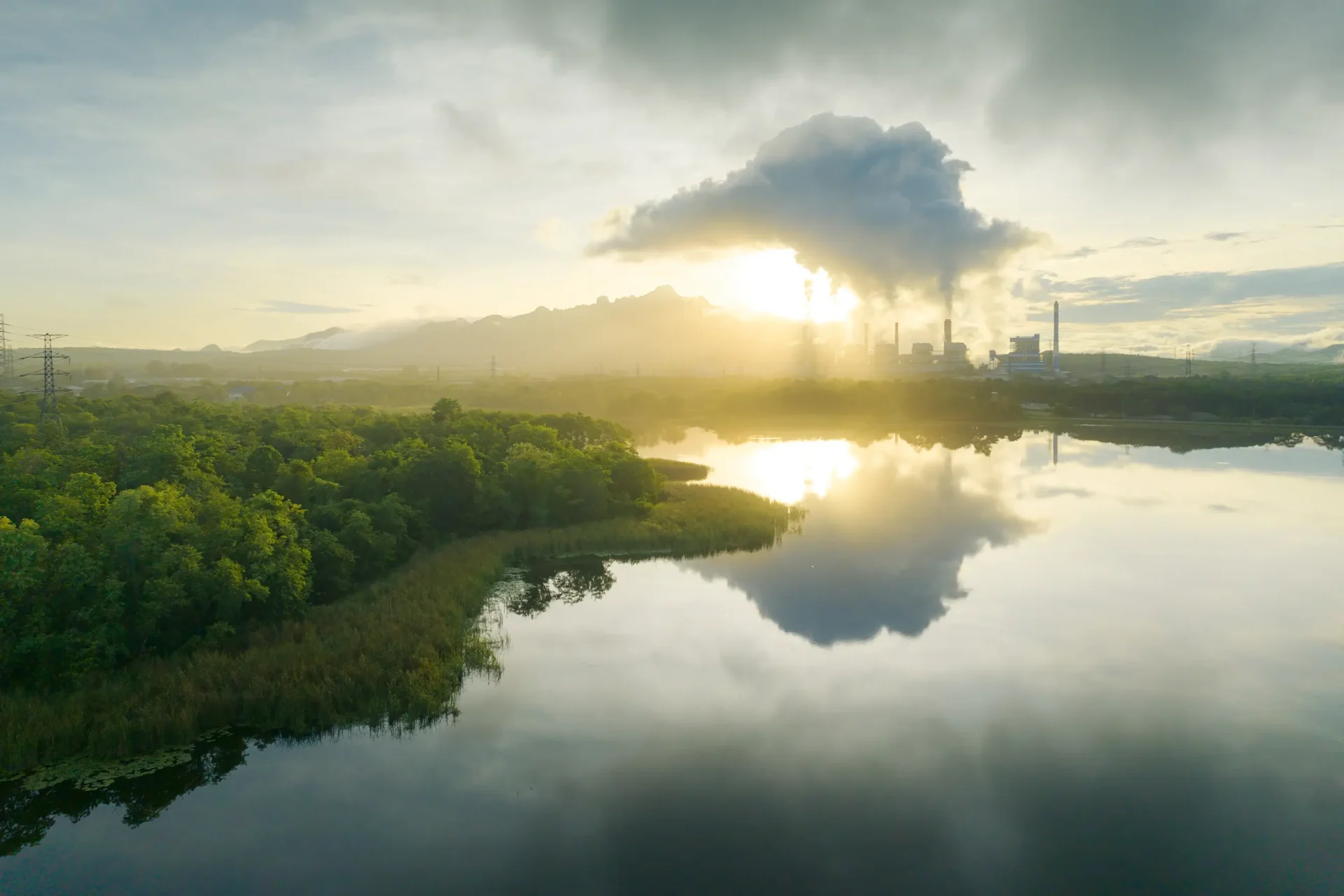 A factory with smokestacks emits clouds near a calm lake surrounded by greenery. As the sun sets behind distant mountains, it offers a contemplative setting perfect for earth research courses studying industrial impact and environmental harmony.