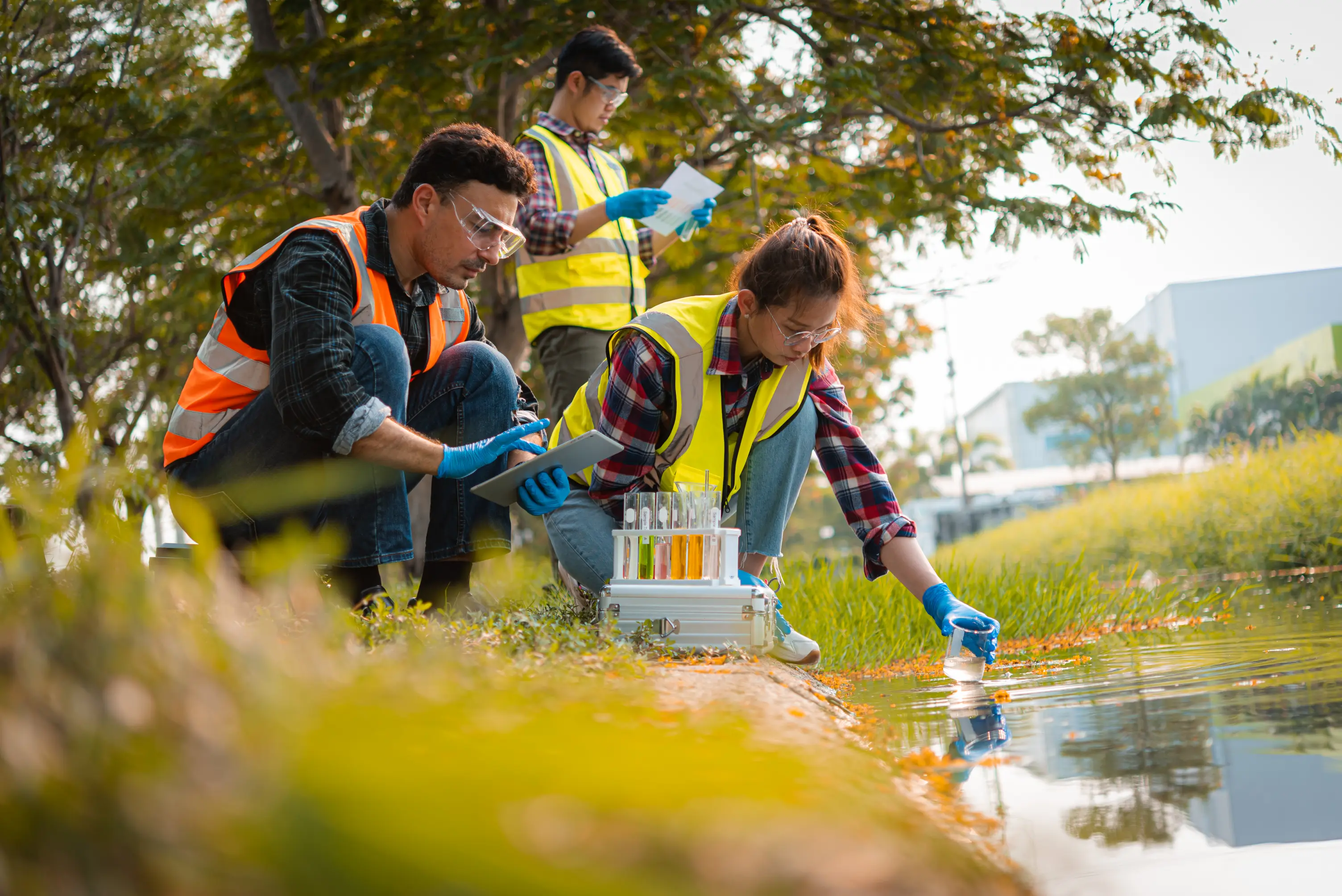 Three researchers in safety vests and goggles, part of an earth research course, collect water samples by a riverbank using test tubes and equipment.