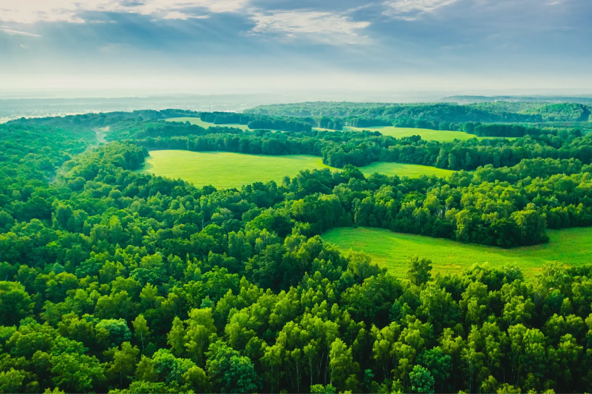 Aerial view of lush green forests and fields under a cloudy blue sky, with sun rays illuminating the landscape.