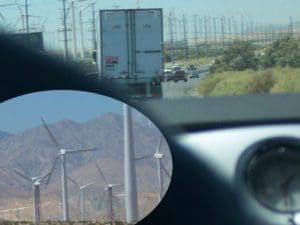 View from a car window showing a highway with a truck and wind turbines in the distance. An inset highlights multiple wind turbines against a mountainous backdrop, sparking curiosity for nature research courses dedicated to renewable energy innovations.